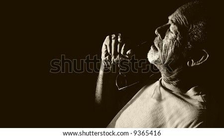 Elderly man with eyeglasses in hand leaning back, thinking. Monochrome B&W portrait on black.