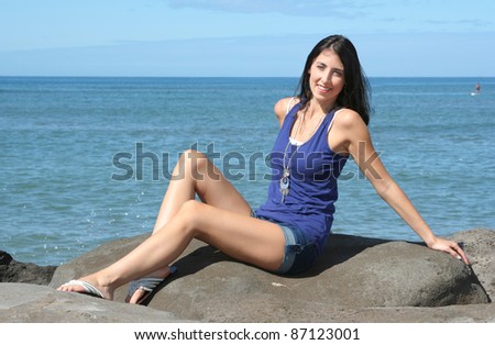 Young attractive woman in blue cotton tanktop and shorts leaning back in the sunlight, on rocks overlooking the blue ocean.