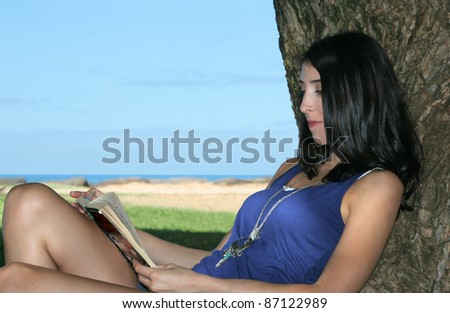 Brunette young woman reading a book in the shade of a tree, overlooking the ocean.