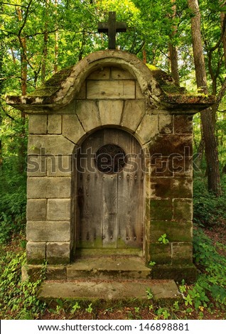 Old traditional village chapel in the park at old graveyard in shadows of old alder trees and lindens. Door with rusty forged gate is closed.