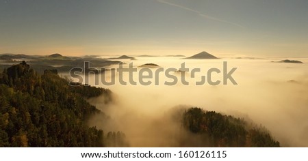 Full moon night with sunrise in a beautiful mountain of Bohemian-Saxony Switzerland. Sandstone peaks and hills increased from foggy background, the fog is orange due to sun rays.