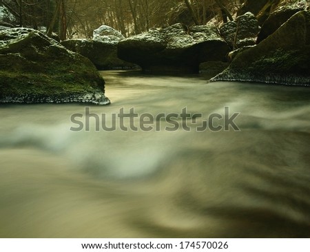 Dark green cold water of mountain stream in winter time between big boulders with snowflakes of first powder snow.