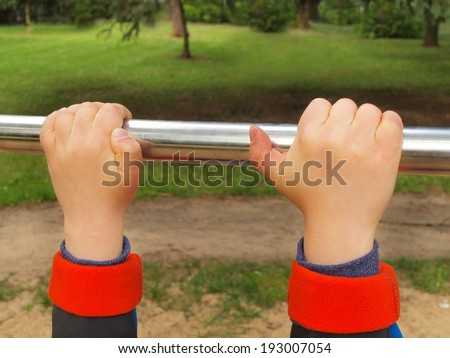Small children hands are hanging on steel horizontal bar. Children playground with green park in the background