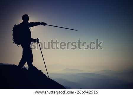 Tourist guide show the right way with pole in hand. Hiker with sporty backpack stand on rocky view point above  misty valley. Sunny spring daybreak in rocky mountains.