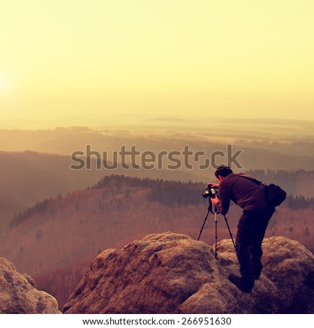 Professional on location and nature photographer  takes photos with mirror camera on peak of rock. Dreamy fogy landscape, spring orange pink misty sunrise in a beautiful valley below.