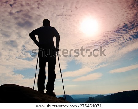 Silhouette of tourist with poles in hands. Sunny colorful daybreak in rocky mountains. Hiker stand on rocky view point above misty valley.