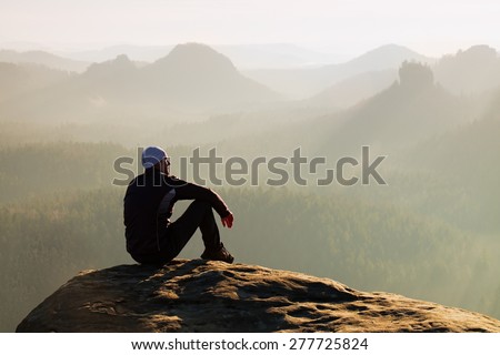 Moment of loneliness. Man sit on the peak of rock and watching into colorful mist and fog in forest valley.