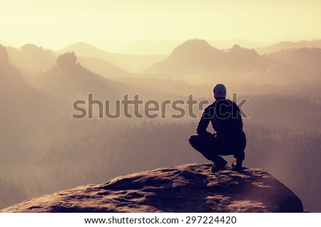 Young man in black sportswear is sitting on cliff\'s edge and looking to misty valley bellow