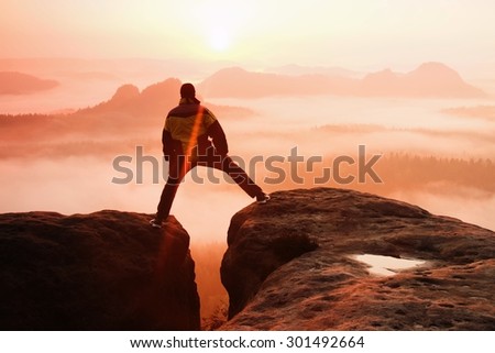Hiker in black is jumping between the rocky peaks. Wonderful daybreak in rocky mountains, heavy orange mist in deep valley.