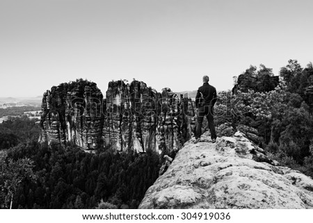 Tall man in black on cliff and watch to mountains. Dark silhouette of rocks. Black and white photo