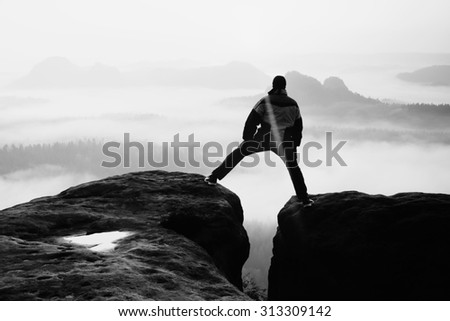 Hiker in black is jumping between the rocky peaks. Wonderful daybreak in rocky mountains, heavy orange mist in deep valley.