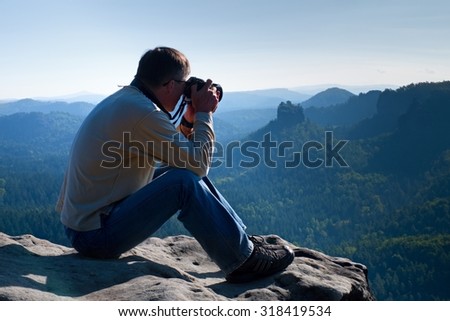 Dark hair man is taking photo by  big mirror camera on the neck on the peak of mountains