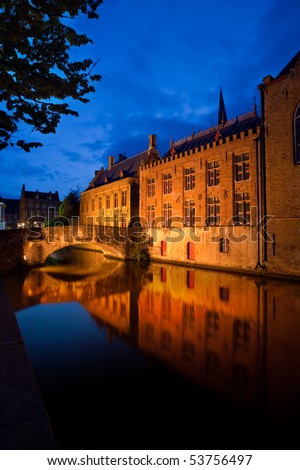 Historic old buildings and a footbridge, illuminated at dusk, are mirrored in a canal.