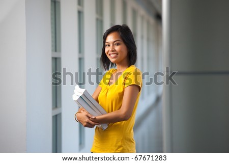 A pretty college student stands smiling with books in a modern hallway on a university campus.  Young female Asian Thai model late teens, early 20s of Chinese descent.