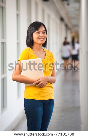 A beautiful singled out Asian girl stands smiling with a book in a hallway on a school campus.  20s female Asian Thai model of Chinese descent.
