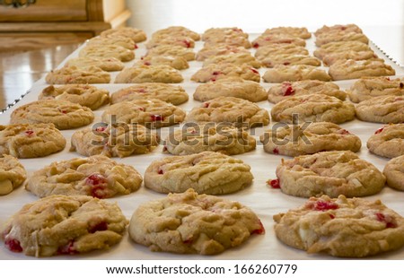 Photo of a batch of fresh baked cookies. They have white chocolate chips with maraschino cherries mixed in.