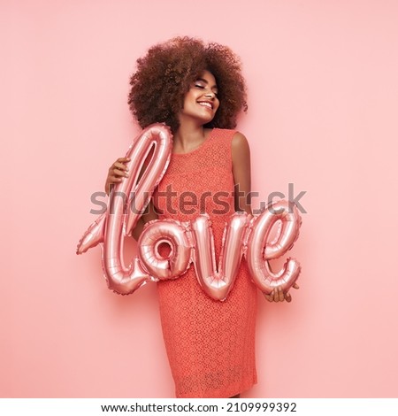 Photo of A beautiful young African-American woman with a balloon in the shape of the inscription love on a pink background. Woman on Valentine's Day. Symbol of love