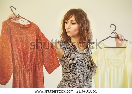 Young woman trying to choose a dress to wear holding up a red and yellow garment on a hanger with a look of indecision as she battles to make up her mind