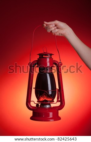 A woman holds up a sooty kerosene lantern. Red backdrop with orange spotlight gives the impression of light emanating from the lantern.