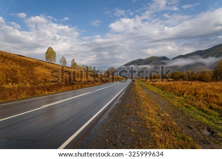Bright picturesque view from the asphalt road, wet after the rain, mountains, fog, golden trees and grass against a blue sky with clouds