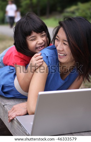 Two young Asian sisters outdoors on a bench with laptop computer laughing happily