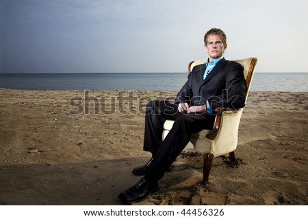 A young man in a suit on the beach with a vintage chair.
