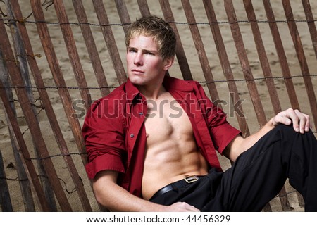 A young muscular man with shirt open leaning on a beach fence.