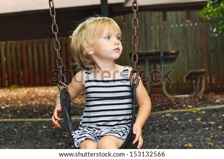 A young blond haired, blue eyed girl looks up from her swing, watching the other children swimg up high thinking about how they do that.