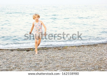 A young, short blond hair, girl, running and playing on a rocky Lake Ontario beach at dusk, at Centennial Park, Hamilton, Ontario, Canada.  Motion visible on hands and arms.