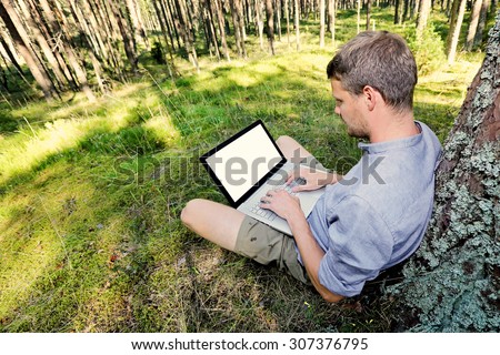 man is sitting against a tree in the forest, working with his laptop