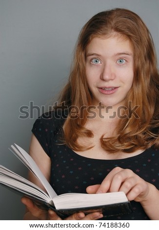 A Young Red Head Girl Points to a Page in an Open Book with a Surprised Expression on her Face, on a Grey Background with Room for Text