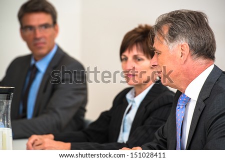 Mixed group in business meeting discussing - focus is on the elderly manager in front and a water bottle on the table