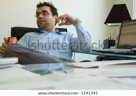 A businessman in a blue shirt sitting at his cluttered desk and looking away.