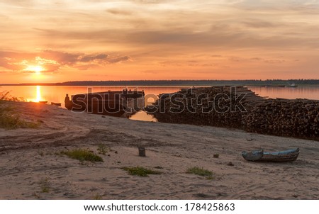 Timber store sinks in water along riverside before cargo ship against panorama of Severnaya Dvina river at sunset .