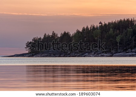 View on Onega Lake and cape with lighthouse among trees against evening glow background at midnight sun. Besov Nos cape, Karelia Republic, Russia