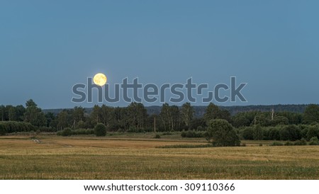 Plain night landscape with fully illuminated orange \'Blue Moon\' (natural phenomenon) over forest with farm field before on August 1, 2015 in  Bulatovo. Kaluzhsky region, Russia.