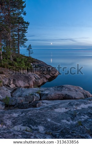 View on Onega Lake granite shore and evening glow with crescent reflecting in calm water at midnight sun. Besov Nos cape, Karelia Republic, Russia.