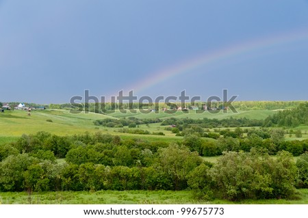 Panorama of hilly landscape with rainbow in the sky against village in background.