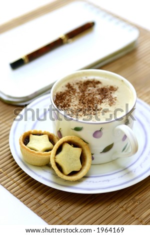 Timeout . A cup of hot cappuccion coffee and mini pies on a bamboo table mat, pen on a notepad in the background.