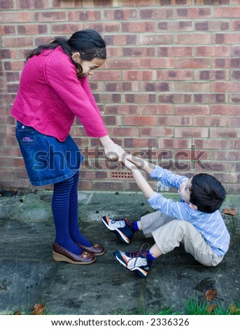 A young girl trying to pull her brother up after he trips over and fall down.