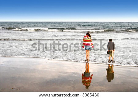 A sister and brother enjoying the waves lapping at their feet as they stood on a sandy beach in a warm evening.
