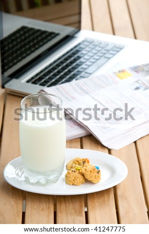 Glass of milk with cookies and early morning newspaper and computer in background.