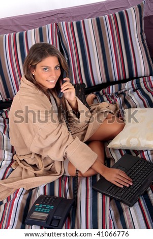 Young attractive woman surfing on internet with laptop on bed in bedroom, while making a phone call.  Studio.