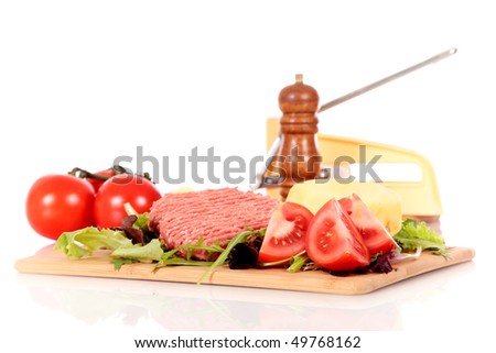 Raw hamburgers, tomatoes and peeled potato on cutting board, French fries cutter and pepper mill on the side,  on reflective surface, studio, white background. Shallow dept, focus on hamburger