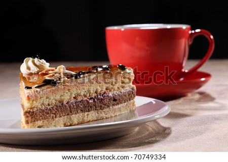 Tasty slice of layered coffee cake on a white plate, along with red coffee cup and saucer. Cake topped with syrup, chocolate sauce, whipped cream and flaked nuts.