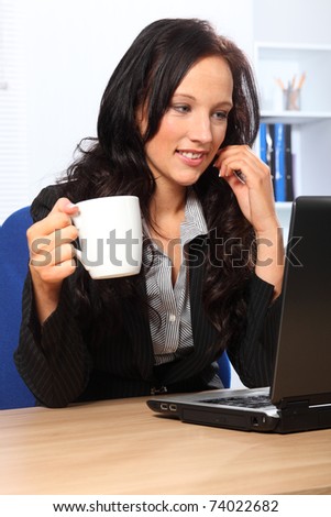 Beautiful young woman enjoying her coffee while working in the office. She has a happy smile and is sitting in front of a laptop wearing a dark business suit.