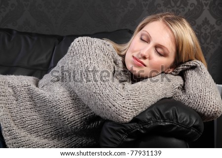 Beautiful young blonde woman lying across black leather sofa at home sleeping, wearing casual grey knitted sweater.