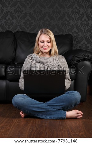 Happy beautiful young blonde girl sitting on the floor at home, bare foot wearing blue jeans and knitted top, surfing the internet on her laptop computer.