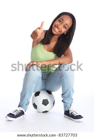 Football success for beautiful young African American teenage school girl soccer player, sitting on a ball with thumbs up sign. Girl wearing blue jeans and casual vest and has a big happy smile.