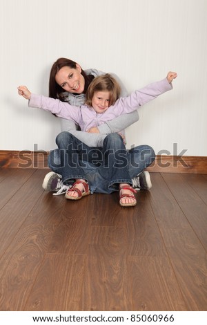 Happy young family of mum and daughter with hug and embrace sitting on the floor at home. Girl has arms raised celebrating success. Both wearing denim jeans and hoodie.